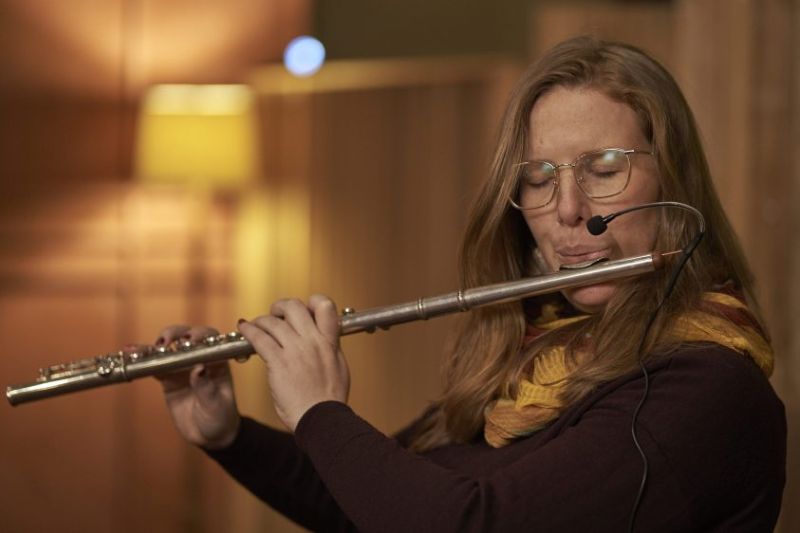 Person with long hair, glasses and eyes closed playa a flute with a microphone attached to it. 