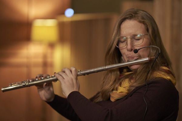 Leah Blankendaal Person with long hair, glasses and eyes closed playa a flute with a microphone attached to it.