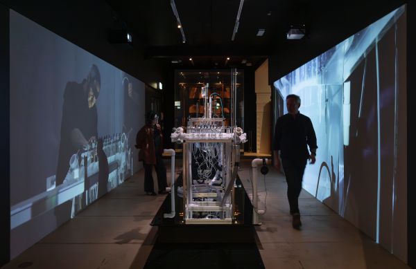two museum visitors look at a sculptural instrument surrounded by two large scale video works