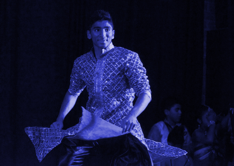 Jainam Mashruwala is cast in a blue light against a dark backdrop. They wear a glittering, intricate robe.