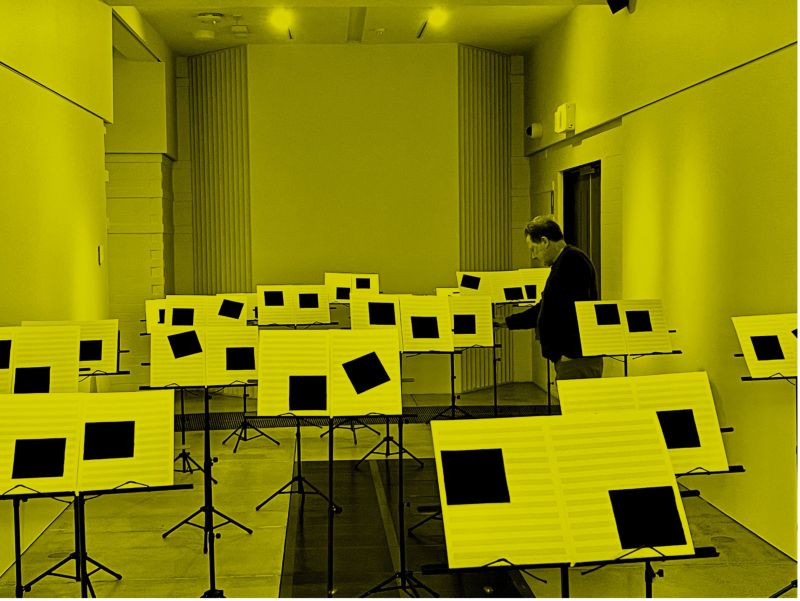 Man standing a room full of music stand with sheet music littered with large black squares, with yellow overlay over photograph